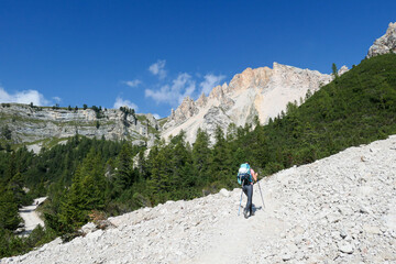 Fototapeta na wymiar A woman with a hiking backpack hiking on a gravelled road in high Italian Dolomites. There is a dence forest at the foothill, and steep and sharp mountain chain in the back. Discovering and exploring