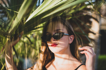 woman wearing sunglasses posing with palm tree. Portrait of beautiful teenage girl with shadows of palm leaf on her face