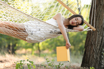 Young woman with book resting in comfortable hammock at green garden