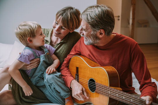 Happy mother and son sitting on bed while father playing guitar in bedroom - Powered by Adobe