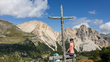 A woman sitting on a wooden bench under a wooden cross end enjoying the panoramic view on a green valley in Italian Dolomites. She is happy and joyful. High and sharp mountains everywhere. Freedom