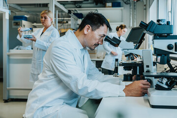 Male scientist sitting by microscope while coworker working in background at laboratory