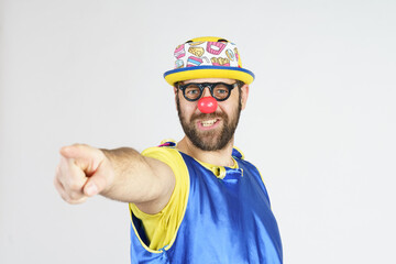 A clown in a bright blue and yellow suit, glasses and a hat, points his finger forward.