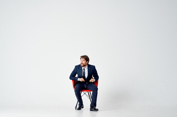 business man sits on red chairs indoors and gestures with his hands on a light background