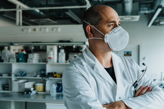 Male scientist wearing face mask looking away while standing with arms crossed at laboratory