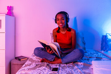 Smiling girl reading book with headphone sitting on bed at home
