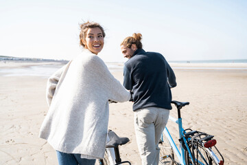 Smiling young woman walking with bicycle and boyfriend while looking over shoulder at beach against clear sky on sunny day