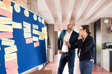 Businessman and woman discussing about note putted on bulletin board while standing at office