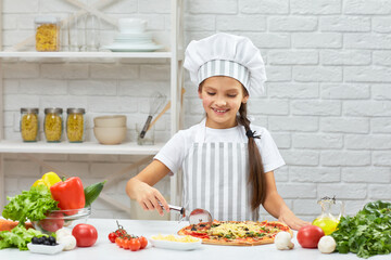 cute smiling little girl in chef hat and an apron cutting pizza at kitchen