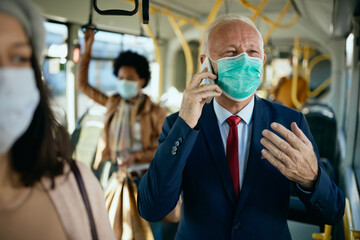 Happy senior businessman with face mask talking on the phone in a public transport.
