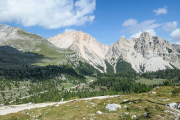A panoramic view on the valley in Italian Dolomites. Dense forest overgrows the mountains slopes. Lush green meadow in front. High mountain chains in the back. Spring in the mountains. Sunny day.