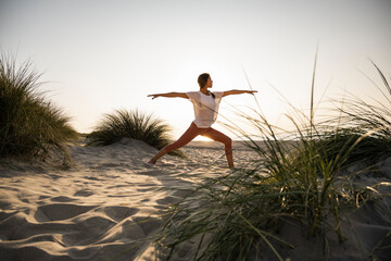 Young woman practicing warrior 2 position yoga amidst plants at beach against clear sky during sunset