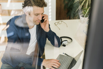 Mid adult man talking on smart phone while using laptop at home