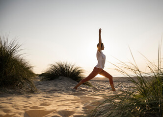 Fototapeta na wymiar Young woman practicing warrior position yoga amidst plants at beach against clear sky during sunset