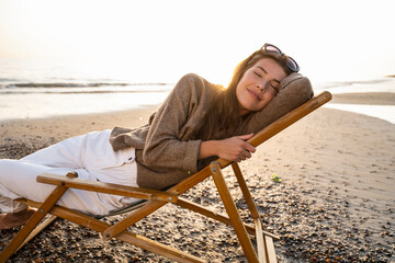 Smiling beautiful young woman reclining on folding chair while relaxing at beach during sunset