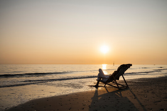 Young woman relaxing while sitting on folding chair at beach against clear sky during sunset