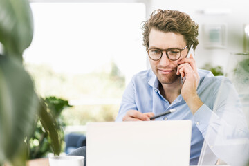Man talking on smart phone while using laptop in office