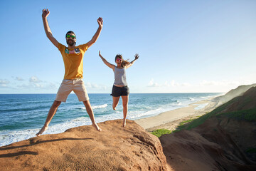 Cheerful couple jumping with arms raised on rock formation at beach