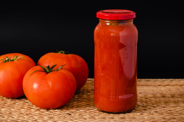 Tres tomates maduros, un frasco de vidrio con tomates triturados, sobre un mantel rústico y fondo negro