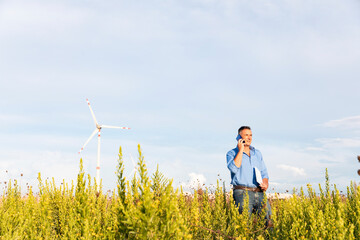 Male engineer talking through mobile phone and document while standing against wind turbines on field