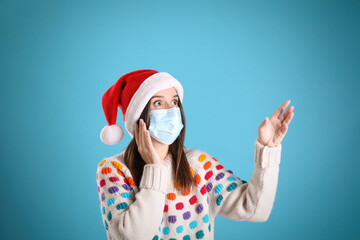 Fototapeta na wymiar Surprised woman in Santa hat and medical mask pointing on light blue background