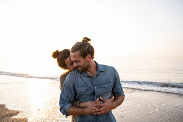 Woman embracing boyfriend from behind at beach against clear sky during sunset