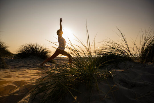 Silhouette young woman practicing warrior position yoga amidst plants at beach against clear sky during sunset