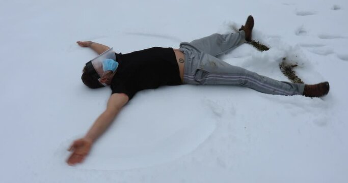 Camera slowly moving around a young man lying in the snow with covid face mask and protective visor, moving arms and legs to make the angel shape.
