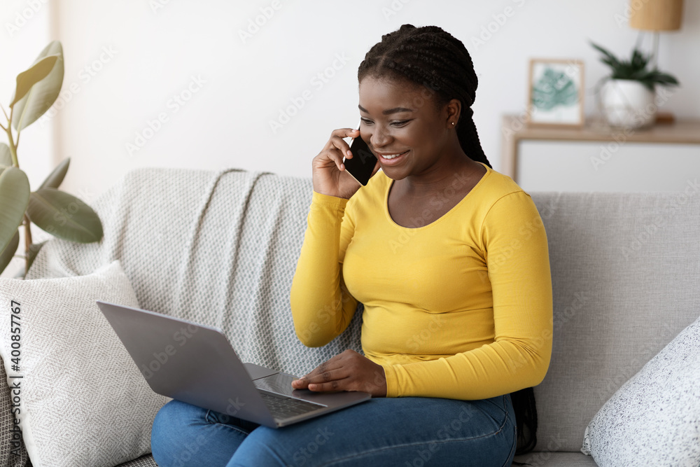 Aufkleber smiling black woman talking on cellphone and using laptop computer at home