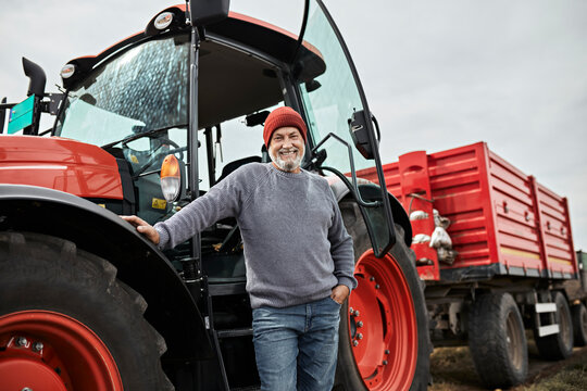Fototapeta Smiling mature farmer standing with hand in pocket against red tractor at farm