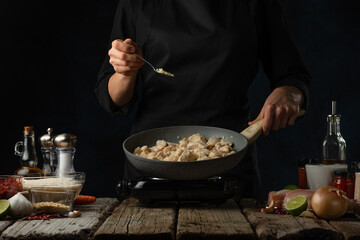 Professional chef pours saffron into pan with frying chicken. Backstage of cooking traditional Indian chicken curry on dark blue background. Frozen motion. Concept cooking process. Frozen motion.