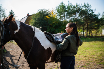 A girl removes a saddle from the back of a horse near a stable on a farm.