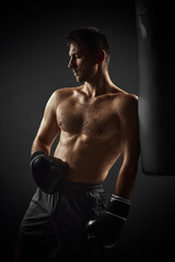 Fototapeta na wymiar Young handsome male boxer in black gloves standing near punching bag on dark background