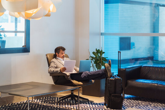 Male professional holding documents while using laptop in hotel lobby