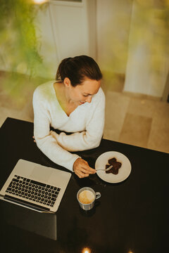 Woman eating chocolate brownie while sitting laptop and coffee at kitchen island