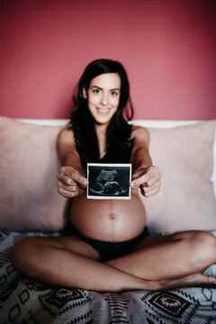 Mid adult pregnant woman showing ultrasound scan while sitting on bed at home