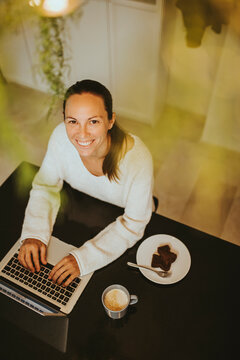 Smiling woman sitting with chocolate brownie and coffee while using laptop at kitchen island