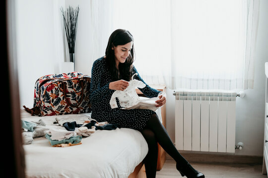 Pregnant woman folding baby clothes in bedroom at home