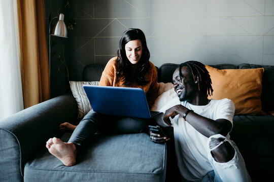 Smiling young man with pregnant woman using laptop while sitting on sofa at home
