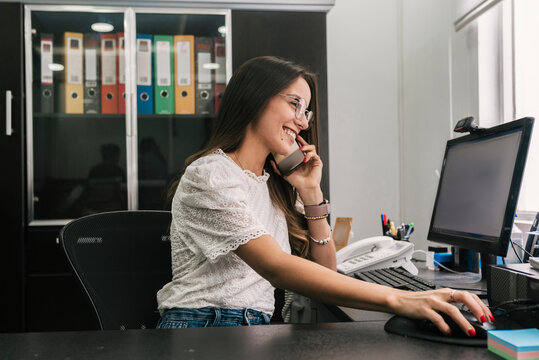 Smiling businesswoman working on computer while talking on mobile phone at office