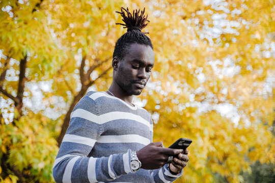 Young man using smart phone while standing against yellow tree in park during autumn