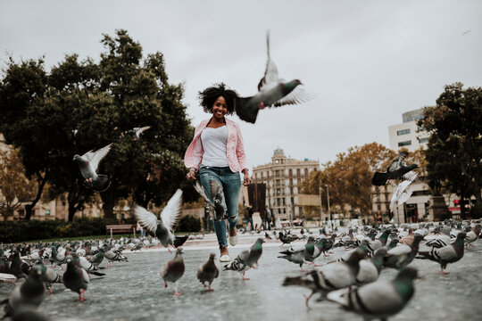 Cheerful woman running amidst flock on pigeons at park in city