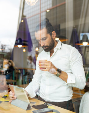 Candid portrait of bearded businessman drinking coffee in front of digital tablet inside cafe