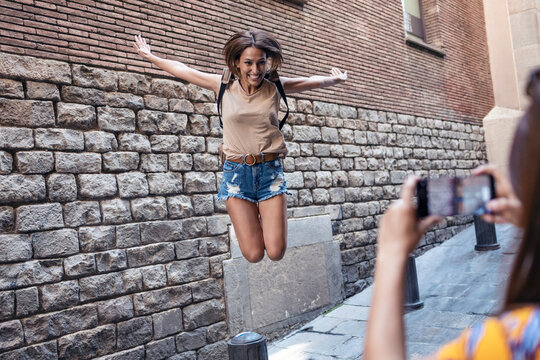 Friend taking photo of woman jumping against wall at Gothic Quarter in Barcelona, Catalonia, Spain
