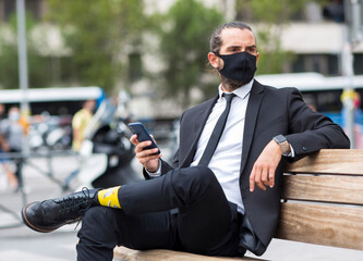 Portrait of businessman wearing protective face mask sitting on bench with smart phone in hand