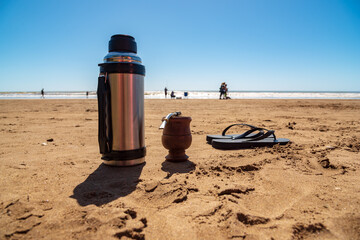 A thermos with a mate and a pair of flip flops resting on the sand on the beach with people walking in the background