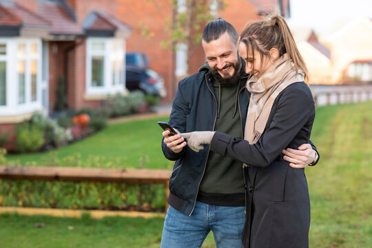 Happy Couple Looking At Smart Phone While Standing In Front Yard