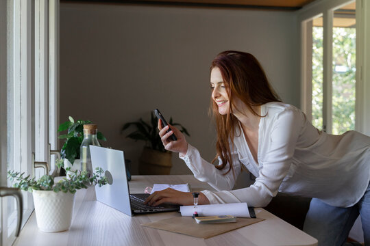 Smiling psychologist using mobile phone while working on laptop at home