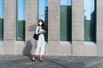 Businesswoman in face mask standing on footpath by building during coronavirus crisis