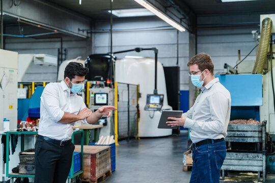 Male coworkers using digital tablet while maintaining social distancing in factory
